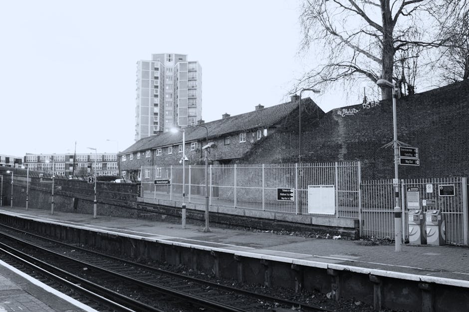 Black and white photograph of Rush Green railway station platform with multiple sets of train tracks in the foreground. The platform is lined with metal railings and features two telephone booths, a w